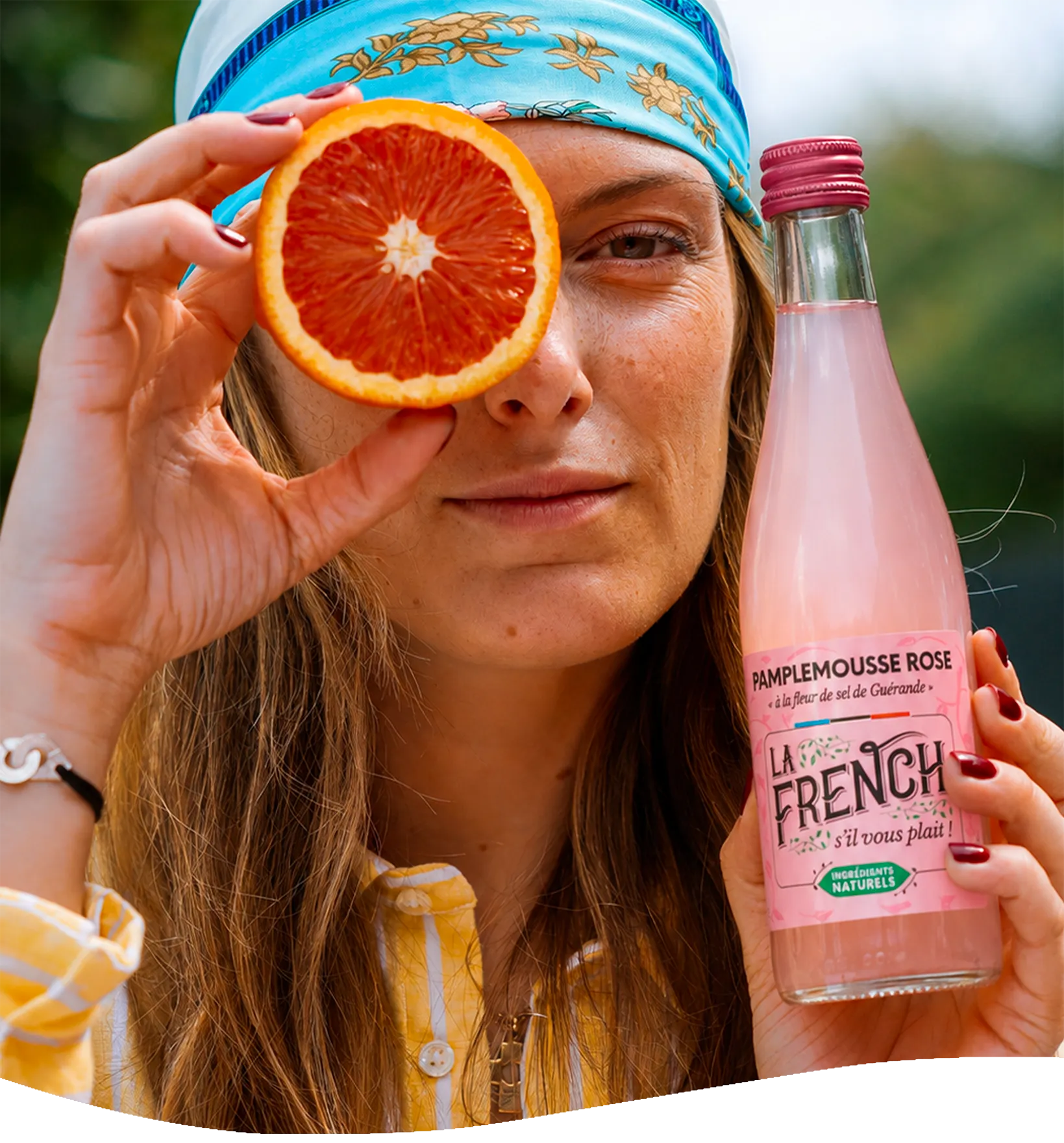 Femme tenant un demi-pamplemousse rose devant son œil et une bouteille Pamplemousse Rose La French
